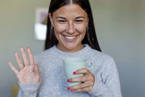 Smiling woman holding a mug and waving during a virtual wellness session or coffee break.